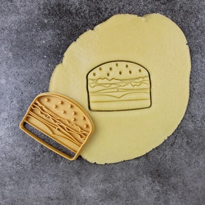 Puede incluir: Un cortador de galletas amarillo en forma de hamburguesa se encuentra sobre una superficie gris junto a una pieza redonda de masa de galletas amarilla. El cortador de galletas se est&aacute; utilizando para cortar la forma de una hamburguesa de la masa de galletas.