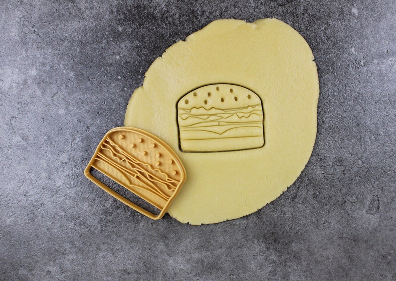 Puede incluir: Un cortador de galletas amarillo en forma de hamburguesa se encuentra sobre una superficie gris junto a una pieza redonda de masa de galletas amarilla. El cortador de galletas se est&aacute; utilizando para cortar la forma de una hamburguesa de la masa de galletas.