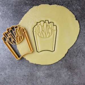 Puede incluir: Un cortador de galletas con forma de caja de patatas fritas. El cortador est&aacute; hecho de metal dorado y est&aacute; sobre una masa de galletas sin hornear.