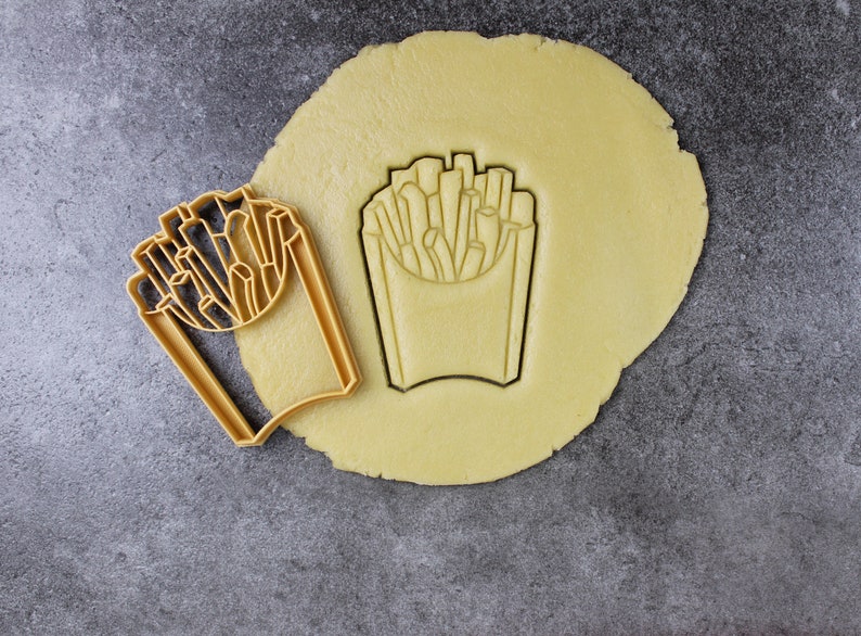 Puede incluir: Un cortador de galletas con forma de caja de patatas fritas. El cortador est&aacute; hecho de metal dorado y est&aacute; sobre una masa de galletas sin hornear.