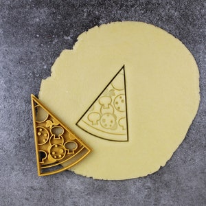 Puede incluir: Un cortador de galletas de color dorado en forma de porci&oacute;n de pizza con ingredientes de pepperoni y champi&ntilde;ones. El cortador est&aacute; encima de un c&iacute;rculo de masa para galletas sin hornear.