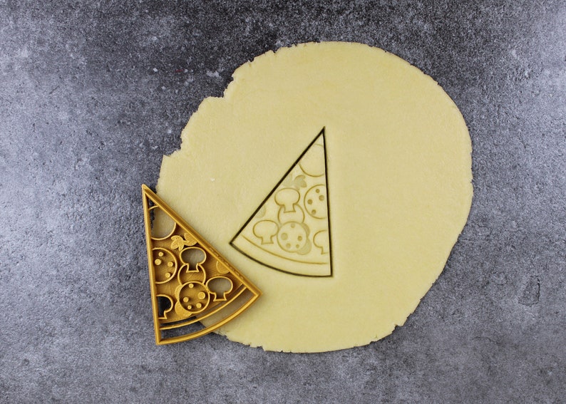 Puede incluir: Un cortador de galletas de color dorado en forma de porci&oacute;n de pizza con ingredientes de pepperoni y champi&ntilde;ones. El cortador est&aacute; encima de un c&iacute;rculo de masa para galletas sin hornear.