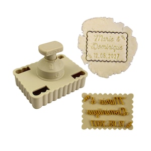 Può includere: Un timbro per biscotti beige con base rettangolare e manico. Il timbro viene utilizzato per imprimere testo sui biscotti. L'immagine mostra anche un biscotto timbrato con la scritta "Marie & Dominique 12.05.2027".