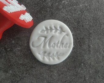 May include: White fondant with the word "Mother" embossed in a cursive font with a leafy design around it. The fondant is being pressed with a red plastic stamp.