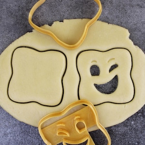 Peut inclure: Gros plan sur une feuille de pâte à biscuits avec deux emporte-pièces dessus. Un emporte-pièce est un visage qui cligne des yeux et l'autre est une forme vide. Les emporte-pièces sont jaunes et la pâte est de couleur brun clair.