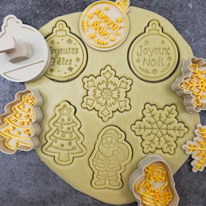 May include: A selection of Christmas-themed cookie cutters. The cutters are beige and yellow, and include shapes such as baubles, snowflakes, Christmas trees, and Santa Claus. The cutters have been used to imprint shapes into a sheet of dough. Some cutters have text that reads "Joyeux Noel" and "Joyeuses Fêtes".