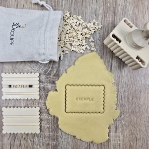 Puede incluir: Un juego de sellos para galletas beige con una bolsa, pequeños bloques de letras y una herramienta de sellado. El juego incluye un corte de galleta con la palabra "EXEMPLE" impresa. La bolsa tiene el texto "3D KOUPE".