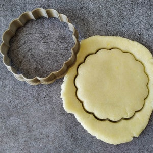 Puede incluir: Primer plano de un cortador de galletas que se utiliza para cortar una galleta redonda de una hoja de masa. El cortador de galletas está hecho de metal y tiene un borde festoneado. La masa es de color amarillo claro.