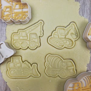 May include: Cookie cutters in the shape of construction vehicles, including a crane truck, excavator, bulldozer, and cement mixer, pressed into dough. The cutters are light beige, and the dough is light yellow.