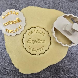 Könnte beinhalten: Ein Keksteig mit einem Ausstecher mit gewelltem Rand und einem passenden Stempel mit Griff. Der Stempel trägt den Namen "Natalia", das Wort "Baptême" und das Datum "16.06.2021". Ein weiterer Stempel trägt den Text "AliaTain Baptême 18.06.2021".