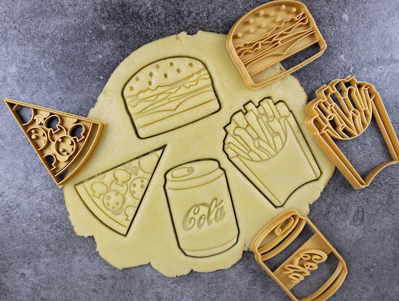 Puede incluir: Un juego de seis cortadores de galletas de color dorado con forma de hamburguesa, patatas fritas, una rebanada de pizza, una lata de cola, una lata de refresco y un recipiente de patatas fritas. Los cortadores est&aacute;n sobre una superficie marr&oacute;n claro.