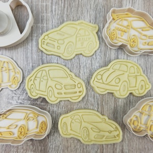 May include: A collection of car-shaped cookie cutters in various designs, including sedans, a sports car, and a classic Beetle. The cutters are light yellow and set against a wooden background. One cutter is a plunger style.