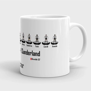 K&ouml;nnte beinhalten: Wei&szlig;e Keramiktasse mit einer Schwarzwei&szlig;illustration einer Fu&szlig;ballmannschaft. Das Team ist Sunderland und die Spieler sind Barton, Gutierrez, Tiote, Carroll und Ameobi. Der Text "Bramble 53" ist in Rot unter der Aufstellung gedruckt.