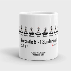 K&ouml;nnte beinhalten: Wei&szlig;e Keramiktasse mit einer Schwarzwei&szlig;illustration einer Fu&szlig;ballmannschaft. Der Text "Newcastle 5 - 1 Sunderland" ist auf der Tasse aufgedruckt, zusammen mit dem Datum "31. Oktober 2010".