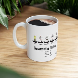 May include: White ceramic coffee mug with a black and white illustration of Newcastle United soccer players. The mug has the team's name and the names of the players printed on it.