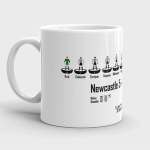 K&ouml;nnte beinhalten: Eine wei&szlig;e Keramiktasse mit einer Schwarzwei&szlig;illustration einer Fu&szlig;ballmannschaft. Die Tasse zeigt die Namen Krul, Coloccini, Enrique, Simpson und Williamson. Der Text "Newcastle 5" ist unter der Aufstellung gedruckt. Der Text "Nolan 26&deg; 34' 75" und "Ameobi 45 70" ist unter dem Teamnamen gedruckt.