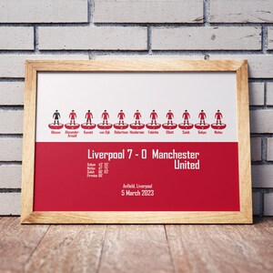 May include: A framed print of a football match lineup with the score Liverpool 7-0 Manchester United. The print features stick figure representations of the players in red and black uniforms. The text below the lineup includes the date of the match, March 5, 2023, and the location, Anfield, Liverpool.
