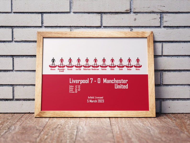 May include: A framed print of a football match lineup with the score Liverpool 7-0 Manchester United. The print features stick figure representations of the players in red and black uniforms. The text below the lineup includes the date of the match, March 5, 2023, and the location, Anfield, Liverpool.