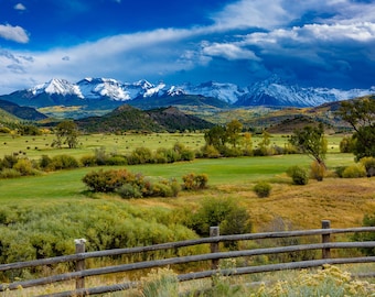 Puede incluir: Una vista panorámica de un prado verde con pacas de heno esparcidas por el campo. Una valla de madera recorre la parte inferior de la imagen. En el fondo, una cordillera con picos cubiertos de nieve se eleva contra un cielo azul con nubes blancas.
