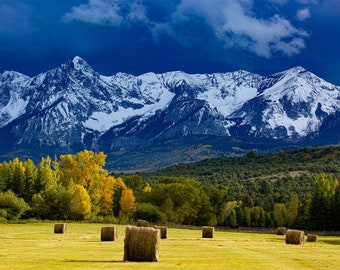 Puede incluir: Un campo de pacas de heno con una cordillera de montañas en el fondo. Las montañas están cubiertas de nieve y el cielo es azul con nubes. El campo es verde y las pacas de heno son marrones.