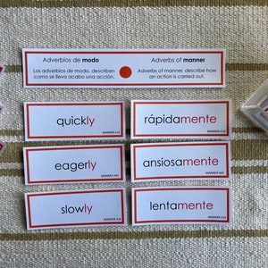 May include: Six white flashcards with red borders, each featuring an English word and its Spanish translation. The words are "quickly", "eagerly", "slowly", "rapidamente", "ansiosamente", and "lentamente". The cards are arranged in two rows of three.