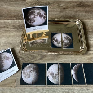 Pode incluir: Um conjunto de oito cartões que mostram as diferentes fases da lua. Os cartões são etiquetados em espanhol com os nomes das fases da lua. Os cartões estão dispostos numa bandeja de prata com um pequeno cavalete de madeira que segura um cartão etiquetado "luna llena" (lua cheia).