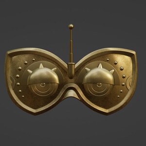 Op de afbeelding: Een goudkleurig masker met twee grote, ronde ogen en een kleine antenne bovenop. Het masker heeft een gedetailleerd, ingewikkeld ontwerp met kleine cirkels en stekels.