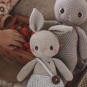 May include: Two handmade crochet bunny dolls. The front doll wears light blue shorts and a strap with a wooden button. The bunnies are light gray with pink cheeks and black eyes. A basket of strawberries is in the background.