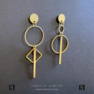 Könnte beinhalten: Ein Paar goldene geometrische Ohrringe. Der linke Ohrring hat einen Kreis, ein Quadrat und einen vertikalen Balken. Der rechte Ohrring hat einen Kreis, einen kleineren Kreis und einen vertikalen Balken.