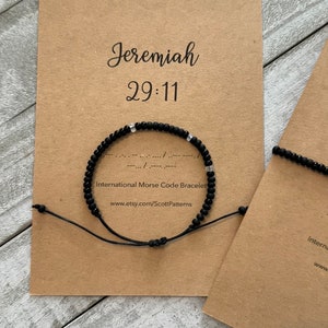 May include: Black beaded bracelet with white beads spelling out a message in Morse code. The bracelet is on a brown card with the text "Jeremiah 29:11" and "International Morse Code Bracelet".