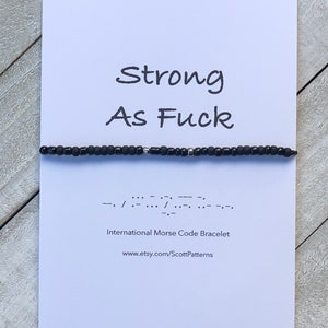 May include: A white card with the text "Strong As Fuck" in black lettering. Below the text is a black beaded bracelet with silver beads. The bracelet is a Morse code bracelet with the code "--/.-..-.--/.-..-.--" printed below it. The text "International Morse Code Bracelet" and "www.etsy.com/ScottPatterns" are printed below the code.