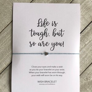 May include: A white card with the text "Life is tough. but so are you!" and a blue string with a silver star charm. The card also includes instructions for making a wish on the bracelet and the text "WISH BRACELET" with a website address.