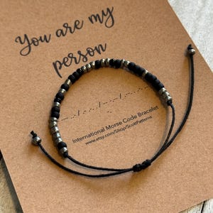 May include: Black beaded bracelet with silver accents, presented on a brown card. The card reads "You are my person" and includes Morse code. The website address etsy.com/Shop/ScottPatterns is also visible.