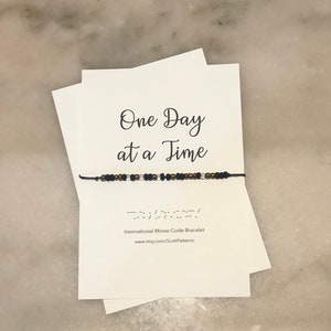 May include: A black and gold beaded bracelet with the phrase "One Day at a Time" written on a white card. The bracelet is made with black and gold beads and a black cord. The card also includes the phrase "International Morse Code Bracelet" and a website address.