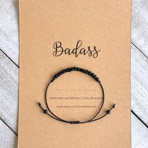 May include: A black beaded bracelet with the word "Badass" spelled out in Morse code. The bracelet is on a brown card with the text "International Morse Code Bracelet" and a website address.