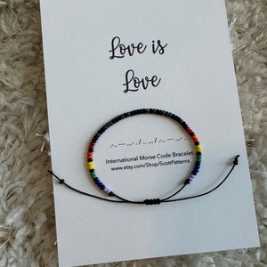 May include: A black string bracelet with rainbow colored beads spelling out "love is love" in International Morse Code. The bracelet is on a white card with the text "Love is Love" and the website address www.etsy.com/Shop/ScottPatterns.