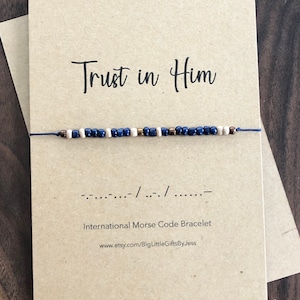 May include: A brown card with the text "Trust in Him" in black cursive font. A blue string bracelet with white, brown, and gold beads is tied around the card. The bracelet is a Morse code bracelet with the code "--- ..- .-.. / .- / .... .--" printed below the bracelet. The text "International Morse Code Bracelet" is printed below the code. The text "www.etsy.com/BigLittleGiftsByJess" is printed below the bracelet description.