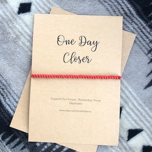 May include: A brown card with the text "One Day Closer" and a red beaded bracelet. The card also includes the text "Support Our Troops - Remember Those Deployed" and a website address.