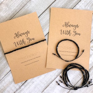 May include: Two cards with the text "Always With You" and black beaded bracelets. The bracelets are made with black beads and black string. The cards are on a light wood surface.