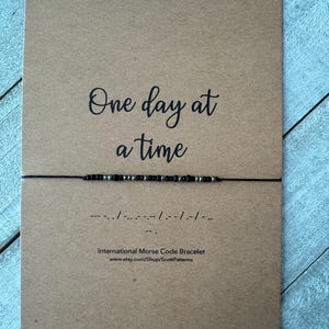 May include: A brown card with the text "One day at a time" in black cursive font. A black string with black and silver beads is attached to the card. The string is tied in a knot and the ends are hanging down. The text "International Morse Code Bracelet" and a website address are printed below the string.