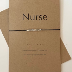 May include: A brown card with the word "Nurse" printed in black. A black string bracelet with silver beads is on top of the card. The bracelet spells out "Nurse" in Morse code. The card also includes the text "International Morse Code Bracelet" and a website address.