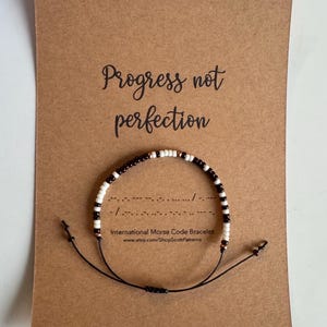 May include: A brown and white beaded bracelet with the message "Progress not perfection" in black text on a brown card. The bracelet is made with black thread and has a sliding knot closure. The card also includes the text "International Morse Code Bracelet" and a website address.