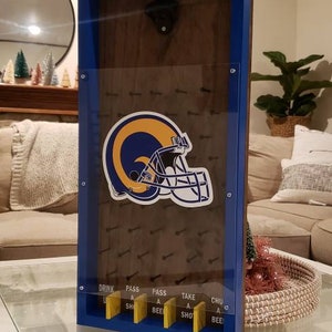 May include: A blue and gold Los Angeles Rams themed bottle opener game. The game features a Rams helmet graphic and a clear plastic panel with a grid of nails. The game has five yellow buttons with the following text: Drink a Beer, Pass a Beer, Take a Shot, Chug a Beer, and Pass a Shot.