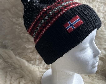 May include: A black knit hat with a red, white, and green patterned band and a black pom-pom. The hat features a Norwegian flag patch. The hat is displayed on a white mannequin head.