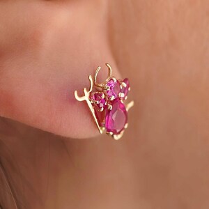 A close-up of a woman's ear showcasing a 14K gold stud earring with a playful pink zirconia devil design, adding a bold and edgy touch. The earring is secured with a screw-back closure, perfect for everyday wear.