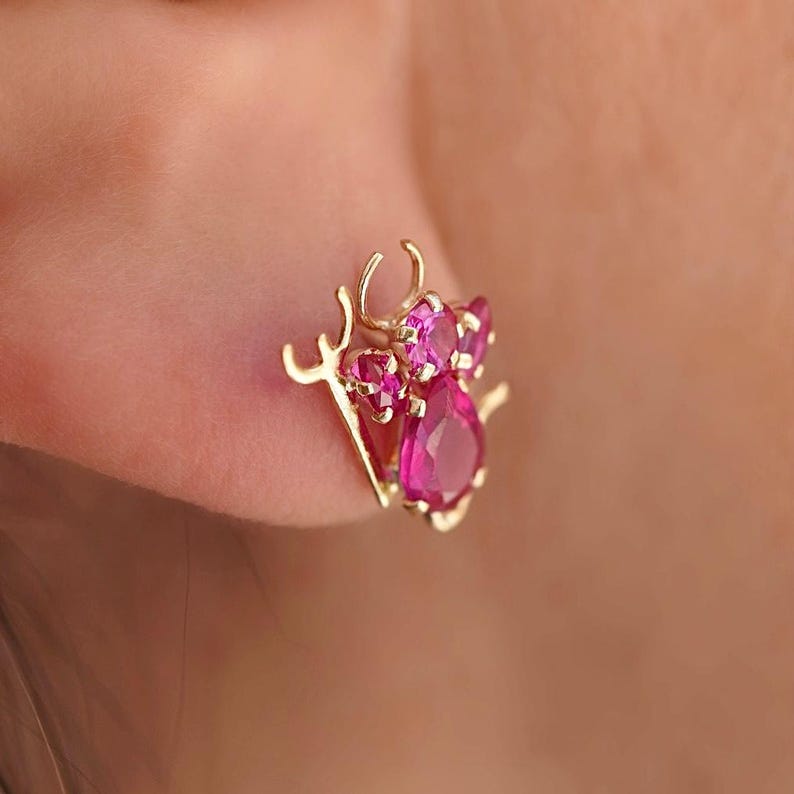 A close-up of a woman's ear showcasing a 14K gold stud earring with a playful pink zirconia devil design, adding a bold and edgy touch. The earring is secured with a screw-back closure, perfect for everyday wear.