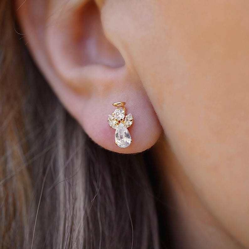 A close-up of a model's ear adorned with a 14K gold stud earring featuring a sparkling white zirconia angel design, symbolizing purity and elegance. The earring is secured with a screw-back closure for a comfortable fit.