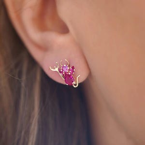 A close-up of a woman's ear showcasing a 14K gold stud earring with a playful pink zirconia devil design, adding a bold and edgy touch. The earring is secured with a screw-back closure, perfect for everyday wear.