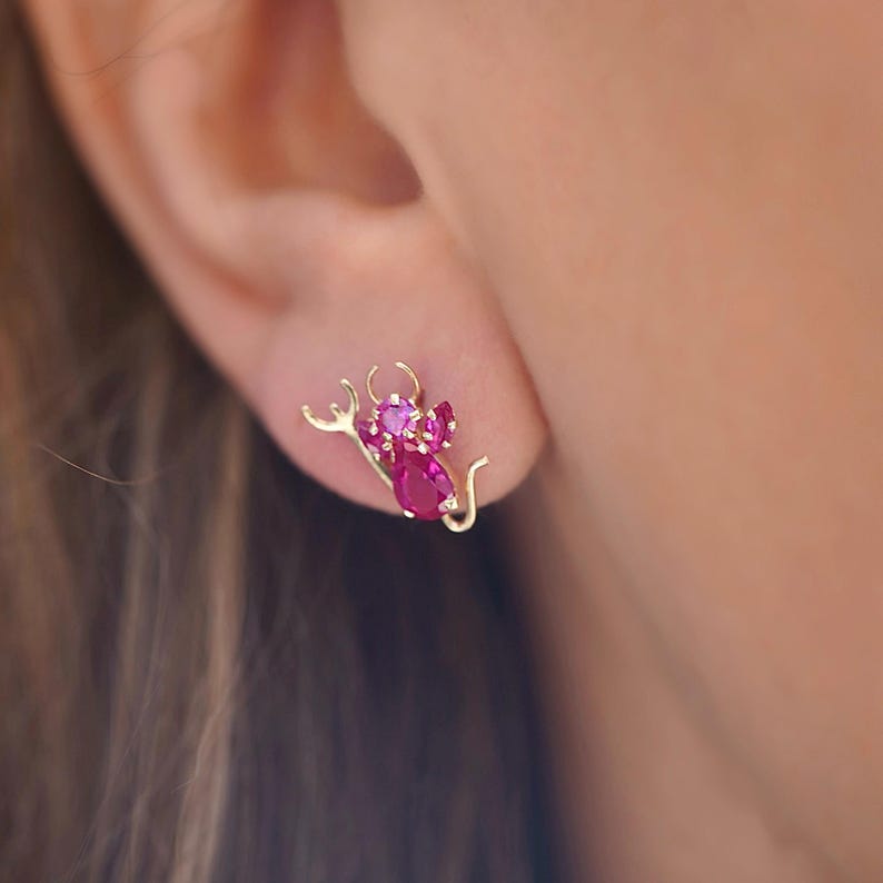 A close-up of a woman's ear showcasing a 14K gold stud earring with a playful pink zirconia devil design, adding a bold and edgy touch. The earring is secured with a screw-back closure, perfect for everyday wear.