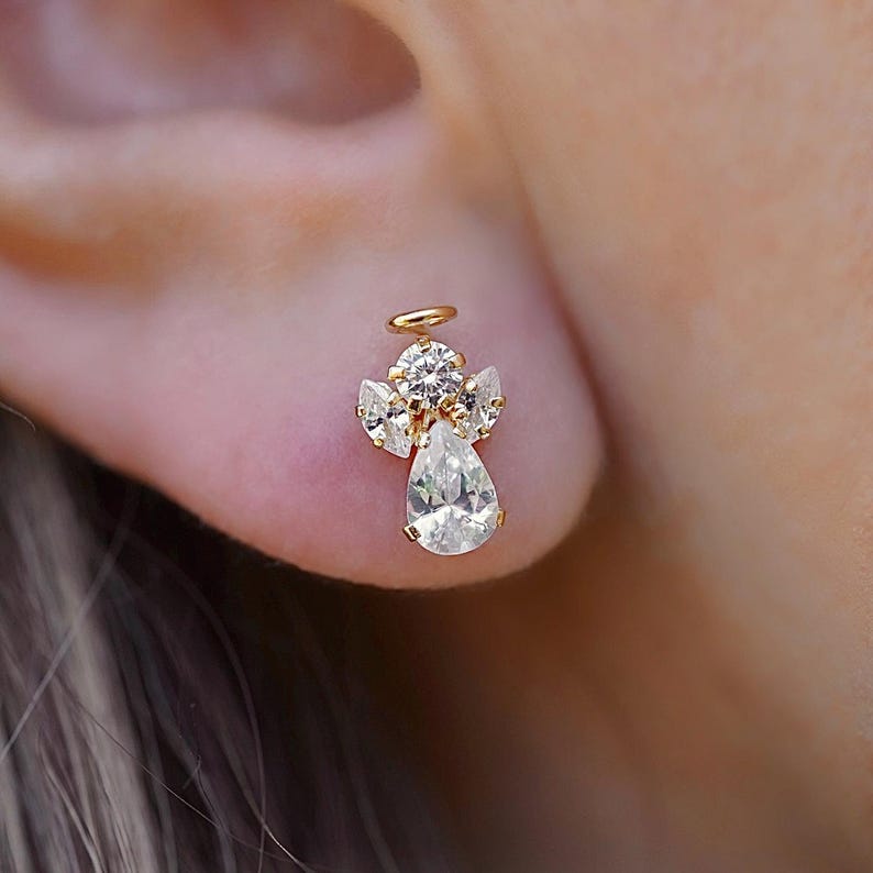 A close-up of a model's ear adorned with a 14K gold stud earring featuring a sparkling white zirconia angel design, symbolizing purity and elegance. The earring is secured with a screw-back closure for a comfortable fit.